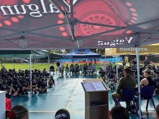 Students seated outdoors under a covered area listening to a speaker at a school assembly
