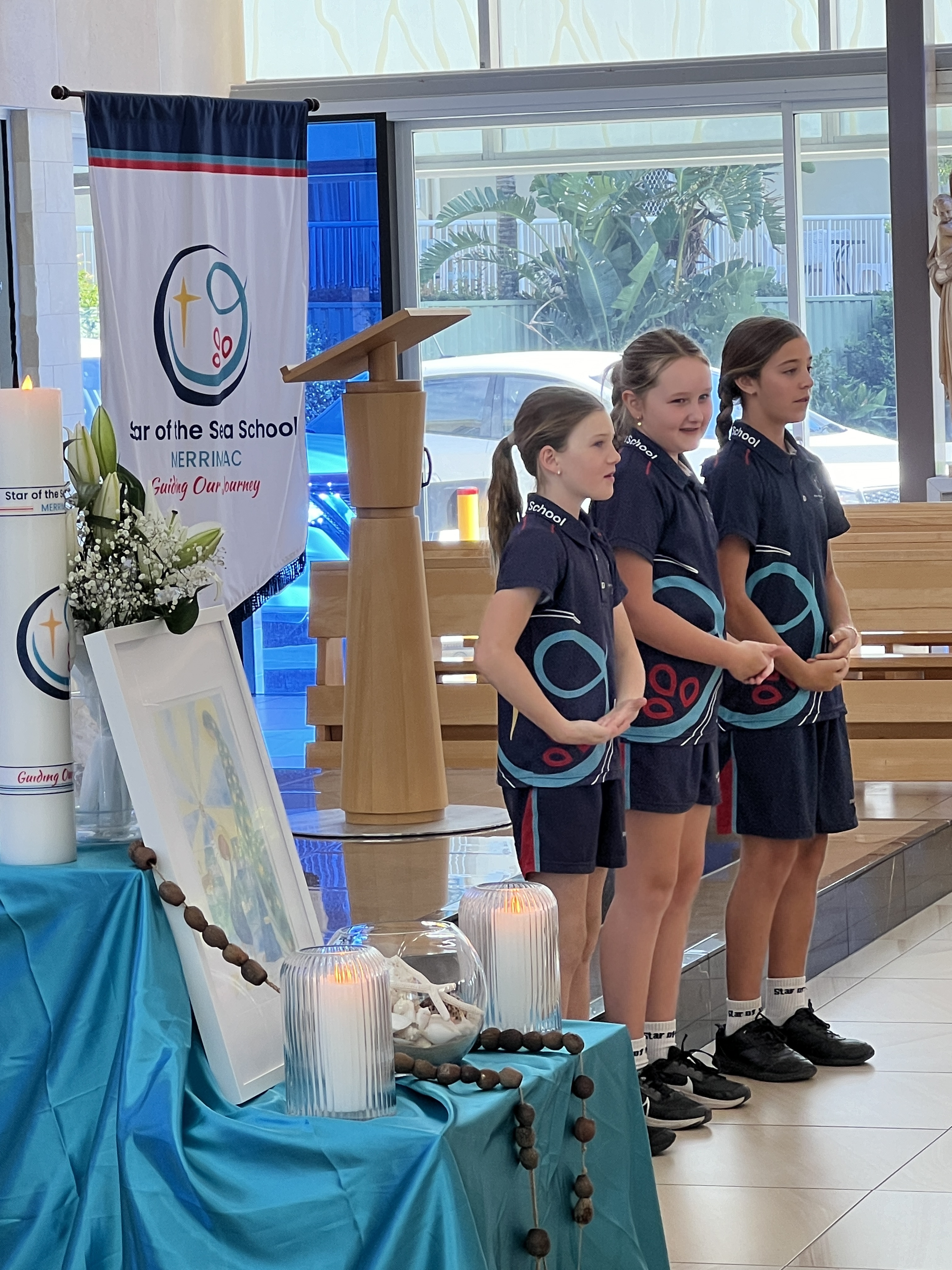 Students standing at the front of a chapel leading a prayer or presentation beside a lectern and school banner