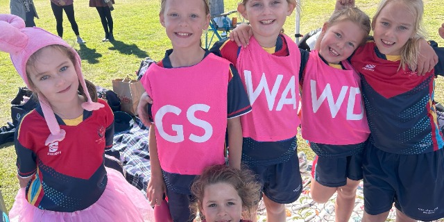 Students in sports uniforms wearing pink bibs standing together on a grassy field during a netball activity