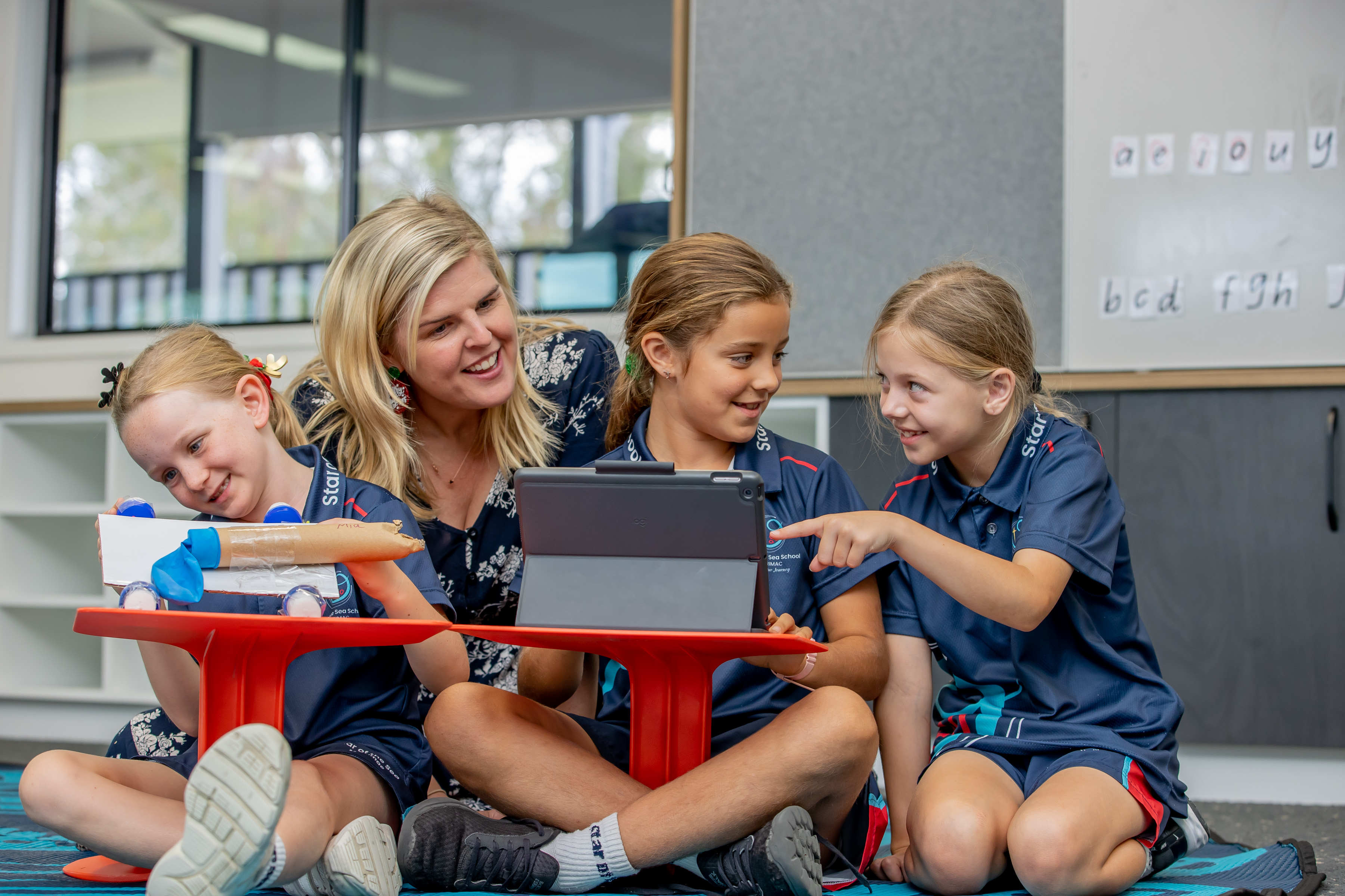 Teacher supporting three students using a tablet while working on a classroom project at small tables