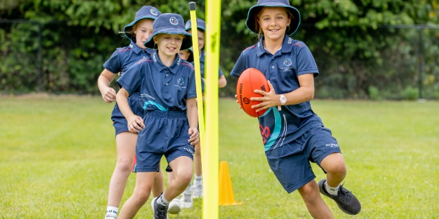 Students playing Australian rules football on a grassy field during a school sports activity.