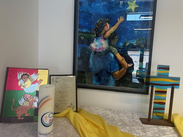 Display with framed artwork, candle, printed text, and colourful cross arranged on a table with yellow cloth